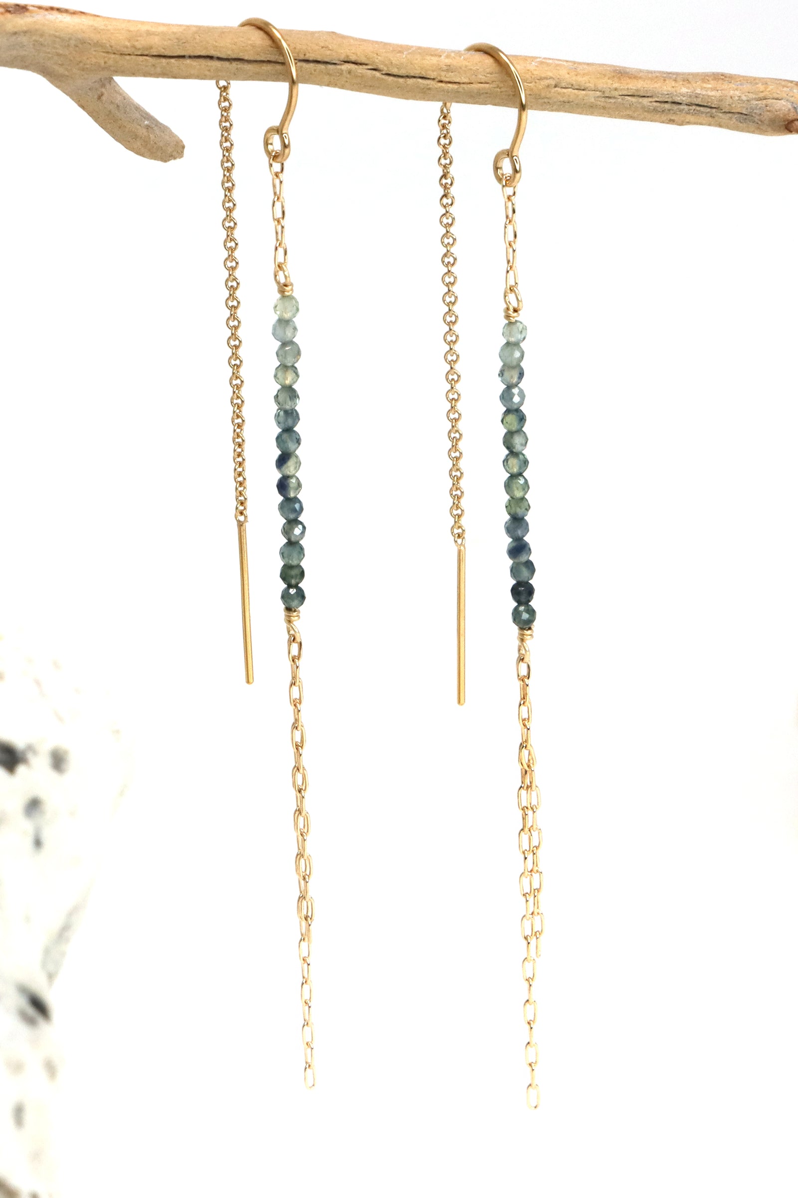 A set of gold-filled threader earrings with faceted sapphire stones, displayed on a wooden branch.